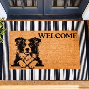 May include: A rectangular doormat with a black and white striped border and a tan center. The center features a black and white illustration of a dog and the word "WELCOME" in black.