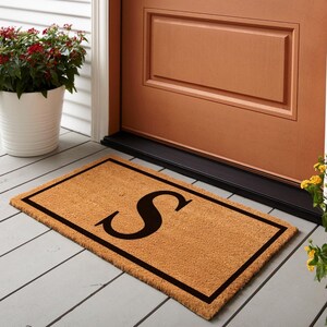 May include: A rectangular doormat made of natural coir fiber, featuring a large black letter "S" in the center. The mat has a black border and is placed in front of a brown door. A white flower pot with red flowers is to the left.