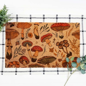 May include: A rectangular doormat with a repeating pattern of various mushrooms and foliage in shades of brown, orange, and red. The mat is made of a natural fiber material and is bordered by a black and white checkered pattern.