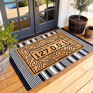 May include: A rectangular tan doormat featuring a black printed circuit board pattern and the text "THERE'S NO PLACE LIKE 127.0.0.1" layered over a larger black and white striped rectangular rug, positioned on a wooden porch in front of a dark gray door.