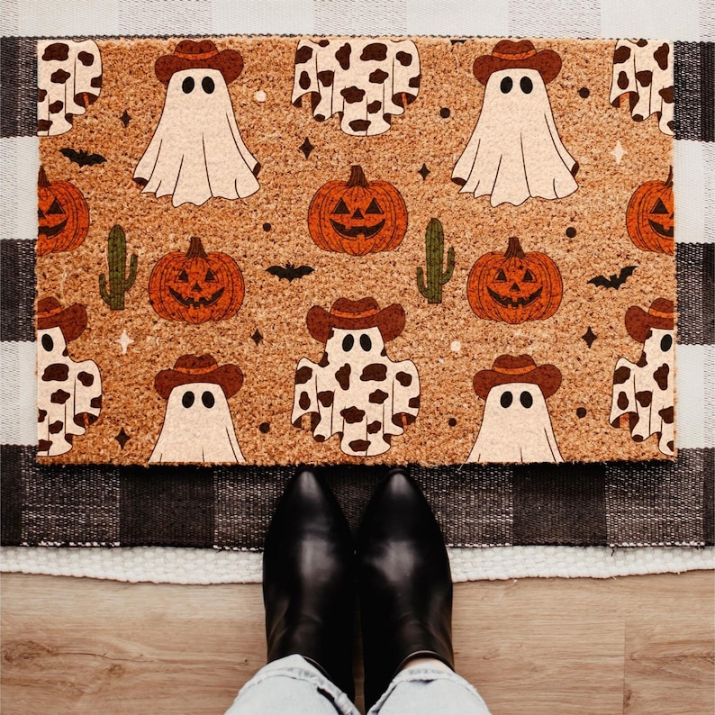May include: A rectangular doormat with a Halloween theme. The mat features a repeating pattern of ghosts wearing cowboy hats, jack-o'-lanterns, bats, and cacti on a brown background. The mat is placed on a black and white checkered rug.