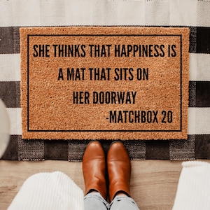 May include: A brown coir doormat with the text "She thinks that happiness is a mat that sits on her doorway - Matchbox 20" printed in black.