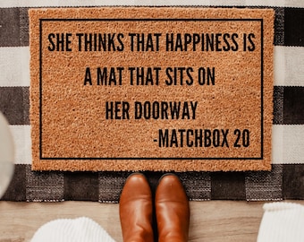 May include: A brown coir doormat with the text "She thinks that happiness is a mat that sits on her doorway - Matchbox 20" printed in black.