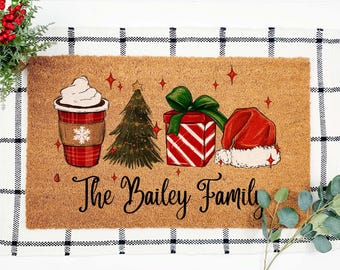 May include: A rectangular doormat with a brown coir surface features Christmas-themed illustrations. A coffee cup, Christmas tree, gift box, and Santa hat are depicted. The text "The Bailey Family" is written in cursive.