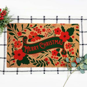 Merry Christmas doormat featuring a dark green holiday banner surrounded by bold red flowers, berries and leafy botanical details on a natural coir-style background. This floral Christmas welcome mat adds rich red and green color to the front porch and creates festive entryway decor for homeowners who prefer botanical holiday styling over traditional character designs.