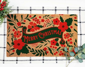 May include: A rectangular doormat with a natural coir fiber base. The mat features a green banner with the words "MERRY CHRISTMAS" in red. Red flowers and green leaves surround the banner, creating a festive holiday design.