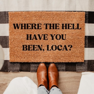 May include: A rectangular doormat made of natural coir fiber, featuring the text "WHERE THE HELL HAVE YOU BEEN, LOCA?" in bold black letters. The mat is placed on a black and white checkered rug, with brown boots and blue jeans visible.