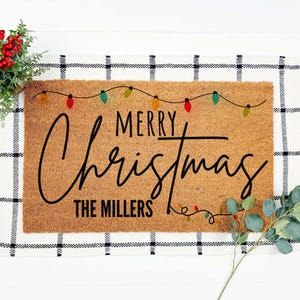 May include: A rectangular coir doormat with the words "MERRY Christmas" in black script, with "THE MILLERS" below. Colorful string lights are above the text. The mat is on a black and white checkered surface.