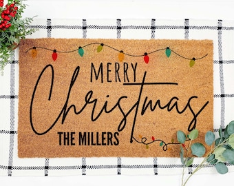 May include: A rectangular coir doormat with the words "MERRY Christmas" in black script, with "THE MILLERS" below. Colorful string lights are above the text. The mat is on a black and white checkered surface.