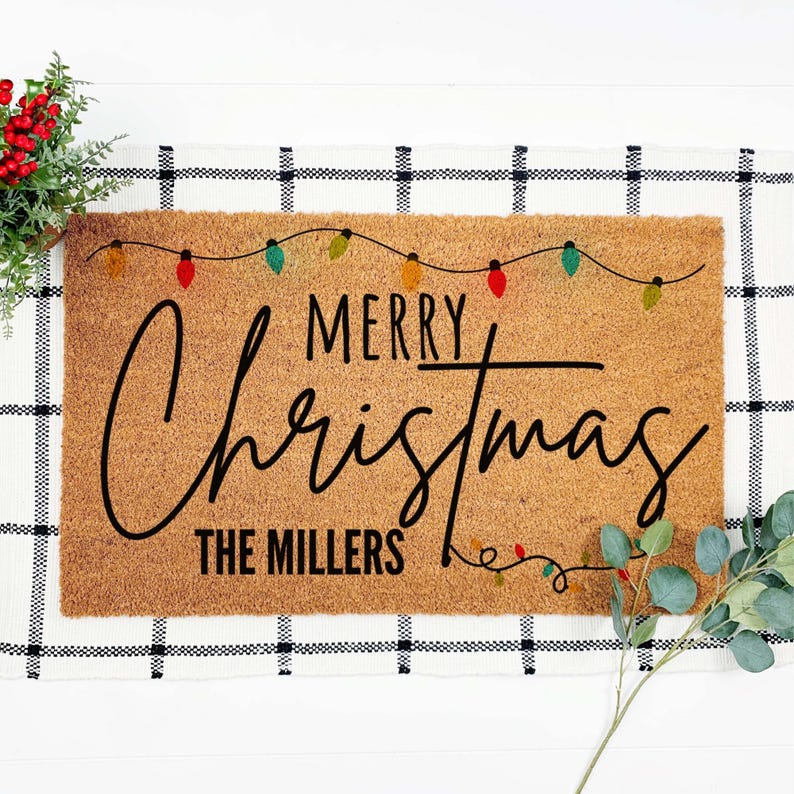 May include: A rectangular coir doormat with the words "MERRY Christmas" in black script, with "THE MILLERS" below. Colorful string lights are above the text. The mat is on a black and white checkered surface.