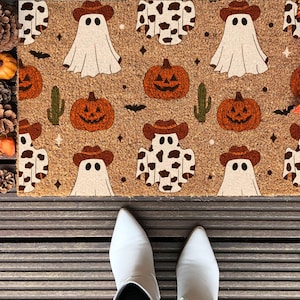 May include: A Halloween-themed doormat featuring a repeating pattern of ghosts in cowboy hats, pumpkins, cacti, and cow-print ghosts. The mat is brown with a textured surface, perfect for seasonal decor. The image also shows white shoes.