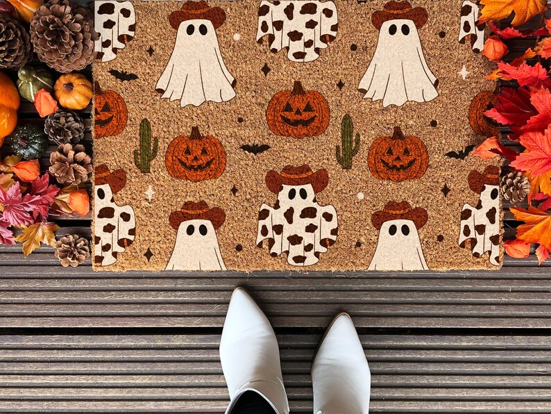 May include: A Halloween-themed doormat featuring a repeating pattern of ghosts in cowboy hats, pumpkins, cacti, and cow-print ghosts. The mat is brown with a textured surface, perfect for seasonal decor. The image also shows white shoes.