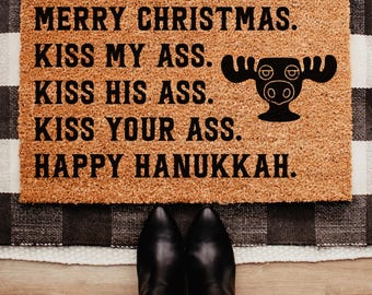 May include: A rectangular doormat with the text "MERRY CHRISTMAS. KISS MY ASS. KISS HIS ASS. KISS YOUR ASS. HAPPY HANUKKAH." in black. A black moose head graphic is on the right. Black boots and blue jeans are visible.