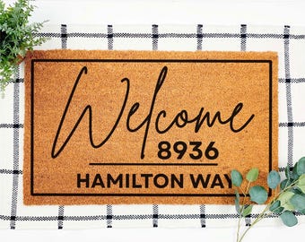 May include: A rectangular doormat with the word "Welcome" in a cursive font, along with the numbers "8936" and the words "HAMILTON WAY" in a block font. The mat is brown with a black border.
