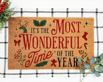 May include: A rectangular coir doormat with the text "It's the Most Wonderful Time of the Year" in red script. The mat features festive illustrations of a reindeer, holly, and a gingerbread man. The background is a white and black checkered pattern.