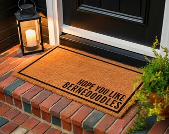 May include: A rectangular doormat with the text "HOPE YOU LIKE BERNEDOODLES" in black. The mat is brown with a black border and is placed on a brick step. A black lantern with a lit candle is to the left.
