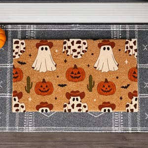 May include: A Halloween-themed doormat with a tan background featuring cartoon ghosts wearing cowboy hats, jack-o'-lanterns, cacti, and bats. The mat is bordered by a gray plaid pattern and sits on a wooden surface.