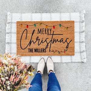 May include: A rectangular doormat with the words "MERRY Christmas" in black script, with "THE MILLERS" below. The mat is brown with a string of colorful lights. It rests on a gray and white plaid rug.