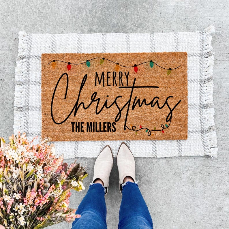 May include: A rectangular doormat with the words "MERRY Christmas" in black script, with "THE MILLERS" below. The mat is brown with a string of colorful lights. It rests on a gray and white plaid rug.