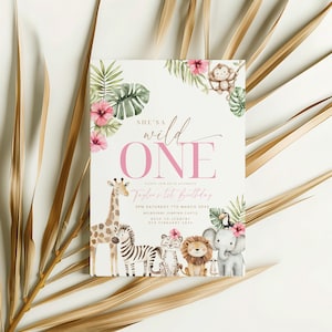 May include: A rectangular invitation card with the text "She's a Wild One" in pink script. The card features watercolour illustrations of a giraffe, zebra, lion, elephant, and monkey, surrounded by tropical leaves and flowers. The card is decorated with a dried palm leaf.