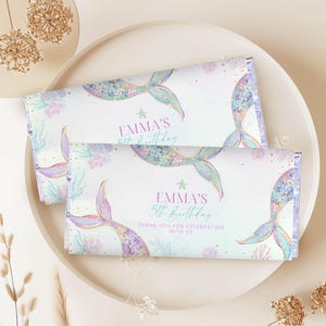 Puede incluir: Dos envoltorios de barra de caramelo con un diseño de cola de sirena. Los envoltorios son blancos con una cola de sirena colorida, coral y el texto "Emma's 5th Birthday". También se incluye el texto "Thank you for celebrating with us".