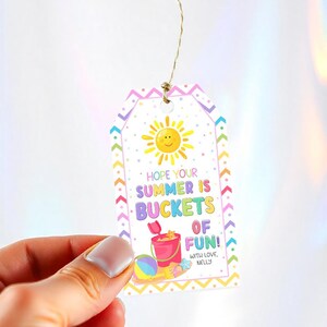 Puede incluir: Una colorida etiqueta de regalo con una ilustraci&oacute;n de sol alegre y el texto "Hope Your Summer Is Buckets of Fun!". Tambi&eacute;n se representa un cubo de playa, una pelota y una estrella de mar. La etiqueta est&aacute; sujeta por una mano.