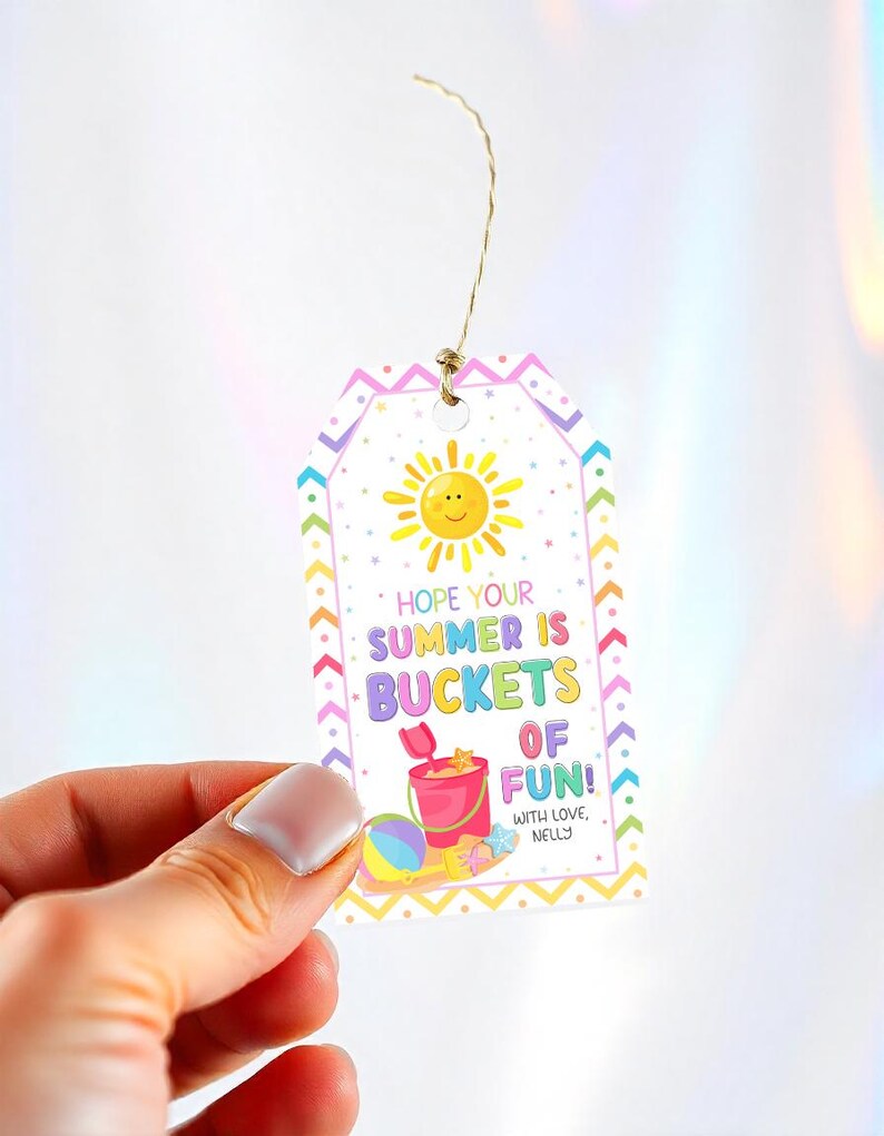 Puede incluir: Una colorida etiqueta de regalo con una ilustraci&oacute;n de sol alegre y el texto "Hope Your Summer Is Buckets of Fun!". Tambi&eacute;n se representa un cubo de playa, una pelota y una estrella de mar. La etiqueta est&aacute; sujeta por una mano.