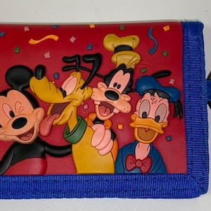 Peut inclure: Portefeuille en tissu rouge et bleu avec un porte-clés. Le portefeuille présente un design 3D de Mickey Mouse, Pluto, Dingo et Donald Duck. Les personnages sourient et sont entourés de confettis.