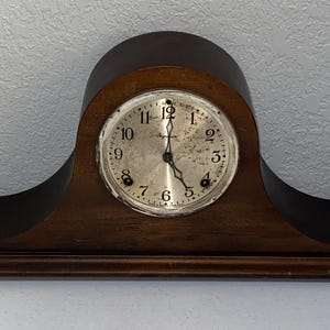Op de afbeelding: Een vintage houten mantelklok met een gebogen ontwerp. De wijzerplaat is zilverkleurig met zwarte cijfers en wijzers. De klok is donkerbruin en staat op een platte basis.