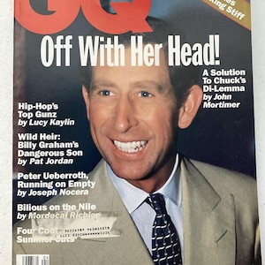 Może przedstawiać: Okładka magazynu GQ z nagłówkiem "Off With Her Head!". Zdjęcie przedstawia uśmiechniętego mężczyznę w garniturze i krawacie. Okładka zawiera tytuły artykułów i czerwono-białe logo. Złoty baner głosi "Regal Clothes For the Working Stiff".
