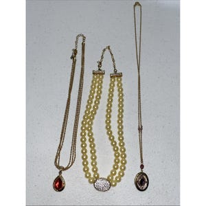 May include: Three gold-tone necklaces: one with a multi-strand chain and a red teardrop pendant, one with two strands of cream-colored faux pearls and a central rhinestone oval, and one with a thin chain and a small oval locket with a dark center.