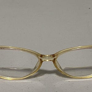 May include: A pair of vintage eyeglasses with a light yellow frame and clear lenses. The frames have a cat-eye shape with brown temples. The glasses are set against a neutral background.