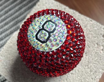 May include: A red, bedazzled billiard ball with the number 8 in the center. The ball is covered in small, red rhinestones, with iridescent rhinestones around the number 8. The number 8 is black with a white background.