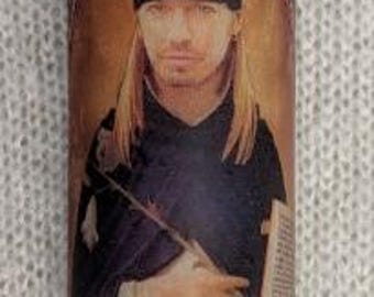 May include: A glass candle with a stained glass design featuring a portrait of a person with long blonde hair wearing a black bandana and a black shirt. The candle is lit and the flame is visible.