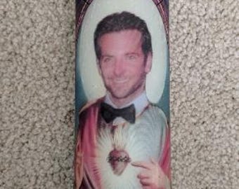 May include: A glass candle with a printed image of a man in a saintly pose, wearing a bow tie and a halo. The candle has a white wax interior and a decorative label with a stained-glass design.