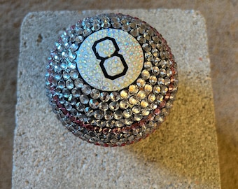 May include: A bedazzled eight ball, a sphere covered in clear and pink rhinestones. The number eight is displayed in a white circle with a black border. The ball is resting on a gray, rectangular block.