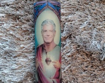 May include: A glass candle with a stained glass design featuring a man with a heart in his chest. The man has short brown hair and is wearing a white shirt. The candle is on a white fuzzy surface.