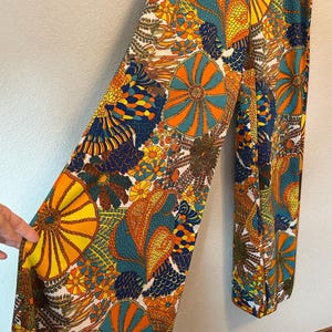 May include: A pair of wide-leg pants with a vibrant, colorful floral pattern. The design features large, stylized flowers and organic shapes in shades of orange, yellow, teal, and navy blue against a white background. The pants are made of a lightweight fabric.