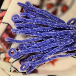 Puede incluir: Un mont&oacute;n de limpiapipas morados, tambi&eacute;n conocidos como tallos de chenilla, est&aacute;n dispuestos en forma de abanico sobre una tela estampada de color blanco y rosa.