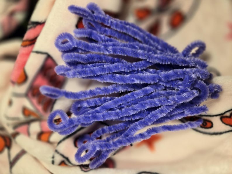 Puede incluir: Un mont&oacute;n de limpiapipas morados, tambi&eacute;n conocidos como tallos de chenilla, est&aacute;n dispuestos en forma de abanico sobre una tela estampada de color blanco y rosa.