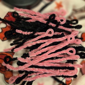 May include: A bunch of pink and black pipe cleaners. The pipe cleaners are twisted together and some are bent into loops.