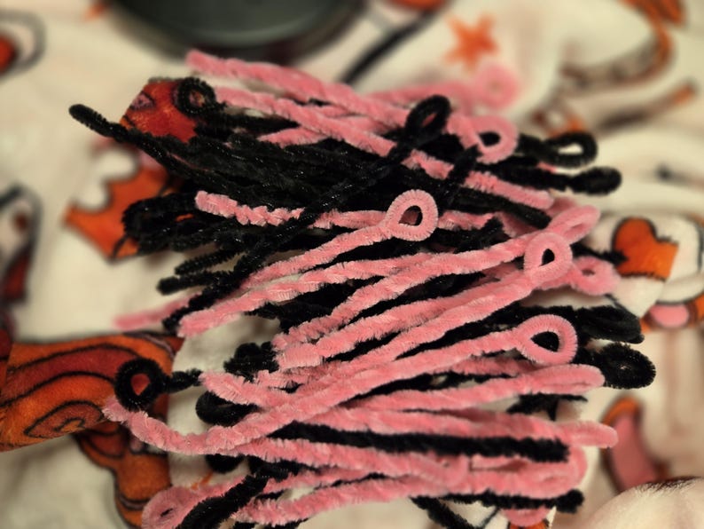 May include: A close-up of a bundle of pink and black pipe cleaners. The pipe cleaners are twisted together and have a fuzzy texture.