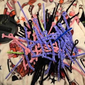 Puede incluir: Un mont&oacute;n de limpiapipas de colores en negro, rosa, morado, azul y verde. Los limpiapipas est&aacute;n dispuestos en un patr&oacute;n aleatorio y est&aacute;n sobre una superficie con estampado blanco y rosa.