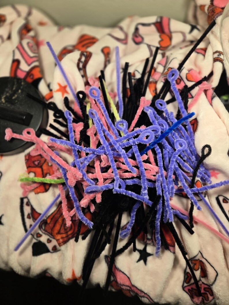 Puede incluir: Un mont&oacute;n de limpiapipas de colores en negro, rosa, morado, azul y verde. Los limpiapipas est&aacute;n dispuestos en un patr&oacute;n aleatorio y est&aacute;n sobre una superficie con estampado blanco y rosa.