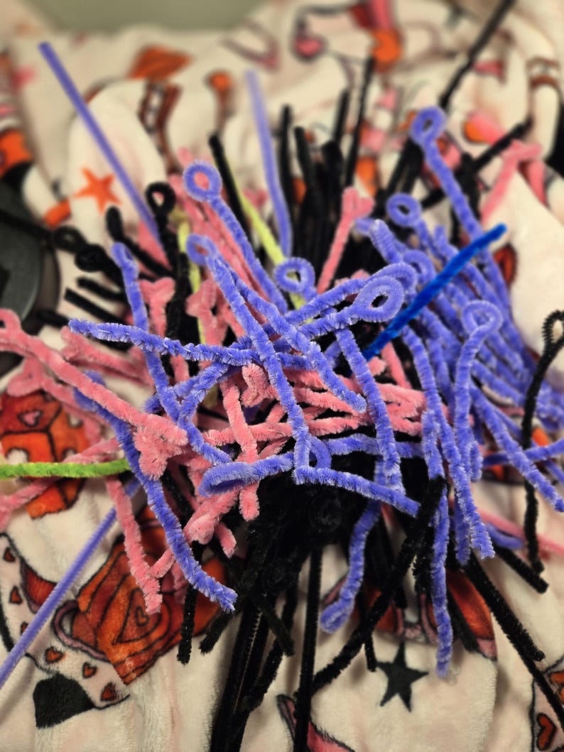 Puede incluir: Un mont&oacute;n de limpiapipas de colores en rosa, morado, negro y azul. Los limpiapipas est&aacute;n dispuestos en un patr&oacute;n aleatorio y est&aacute;n sobre una superficie con un patr&oacute;n blanco y rojo.