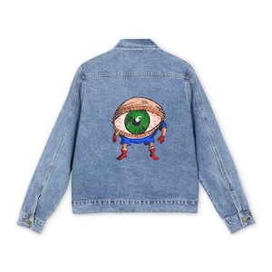 May include: Light blue denim jacket with a colorful cartoon eye graphic on the back. The eye has a green iris, red veins, and is wearing a blue outfit with red boots. The jacket has a classic design with button closures and pockets.