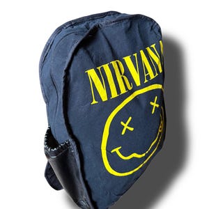 May include: A black fabric backpack featuring the yellow Nirvana band logo and a yellow smiley face graphic with X-shaped eyes. The backpack includes a black side pocket with an elasticized opening and a zipper closure along the top edge.