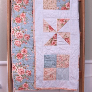 May include: A floral quilt with a light blue background and pink rose pattern. The quilt features a white center with patchwork squares and a pinwheel design. The quilt is displayed on a wooden ladder.
