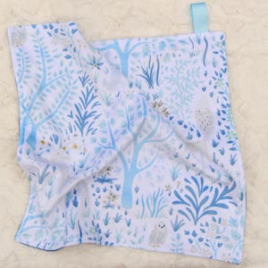 May include: A white baby blanket with a blue and light blue forest pattern featuring trees, foliage, and owls. The blanket has a blue satin ribbon attached to one corner. The blanket is soft and plush.
