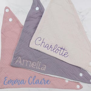 May include: Three triangular bibs with snap closures. The bibs are made of a soft, textured fabric and are embroidered with the names "Emma Claire", "Amelia", and "Charlotte".